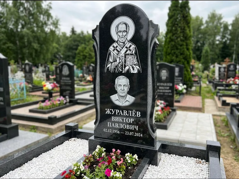 Памятник с гравировкой иконы: стела с образом в полный рост на обороте — гравировка образа Матроны Московской, низкие кусты спиреи в цвету, работа «Лебедева» в Салехарде