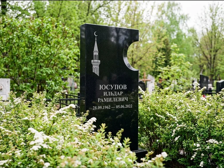 Мусульманский памятник с гравировкой мечети: стела с боковым вырезом в форме полумесяца — гравировка минарета с полумесяцем, низкие кусты спиреи в цвету, работа «Юсупов» в Салехарде